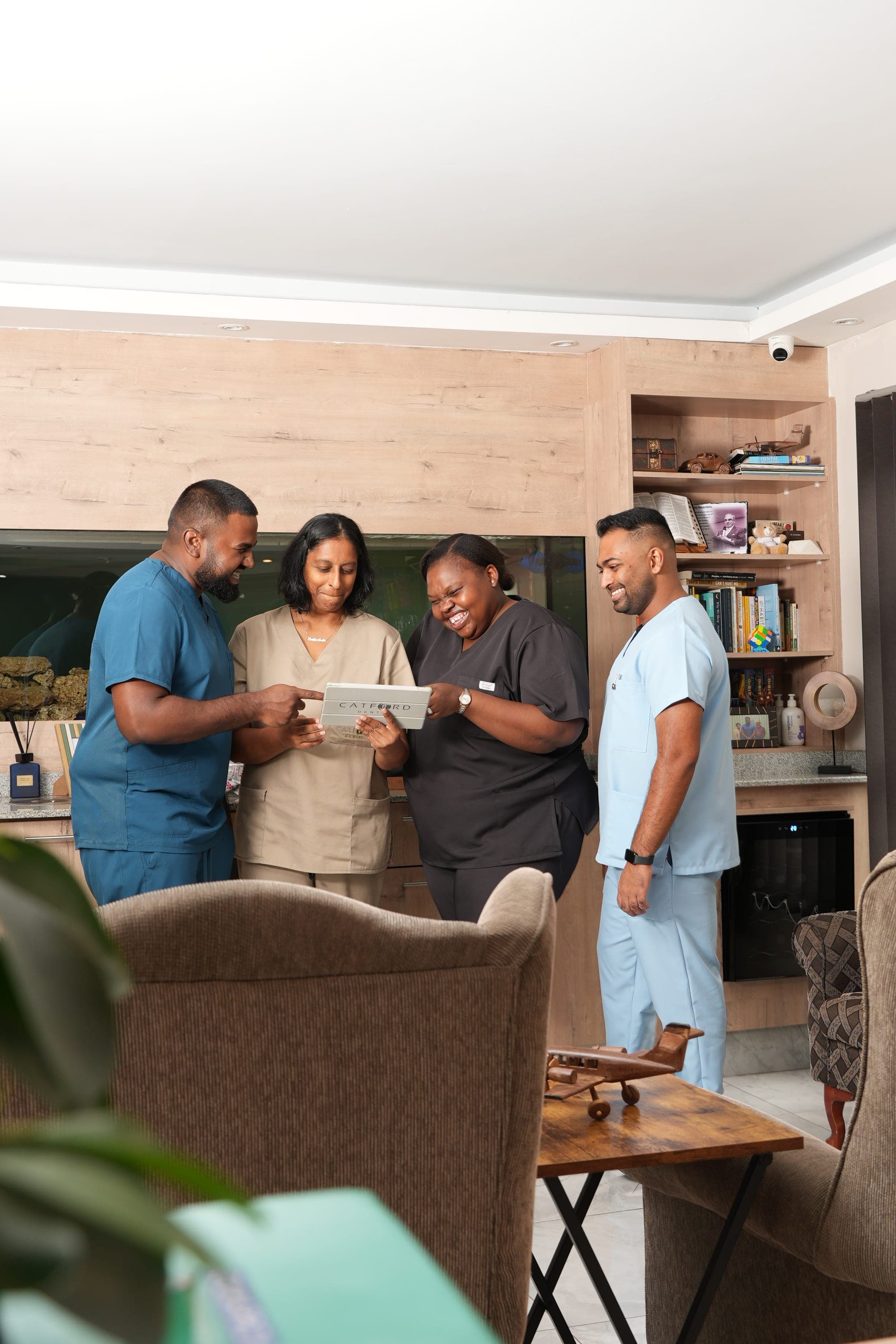 Four members of the Catford Dental team in scrubs, gathered around a tablet and talking, with the practice aquarium behind them.